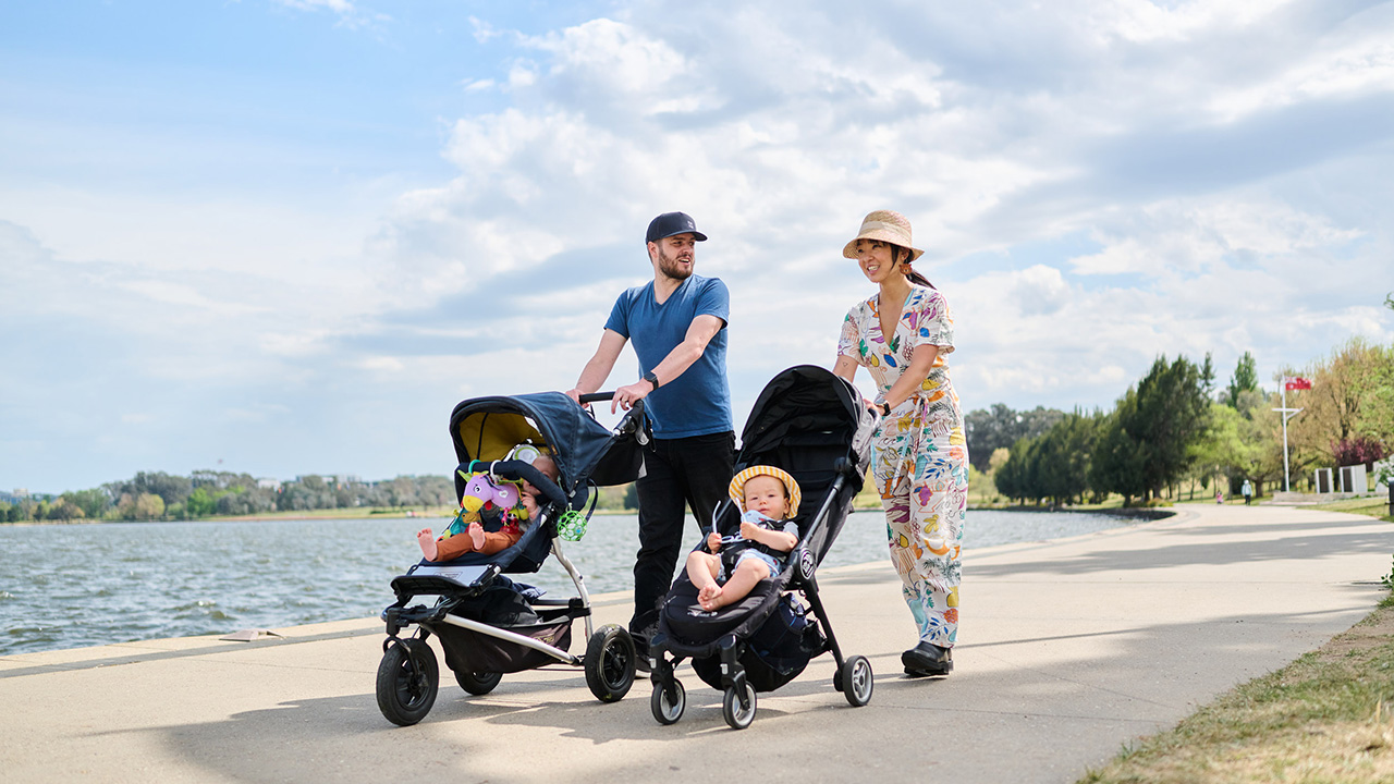 An adult man and woman each push a pram as they walk around a lake.