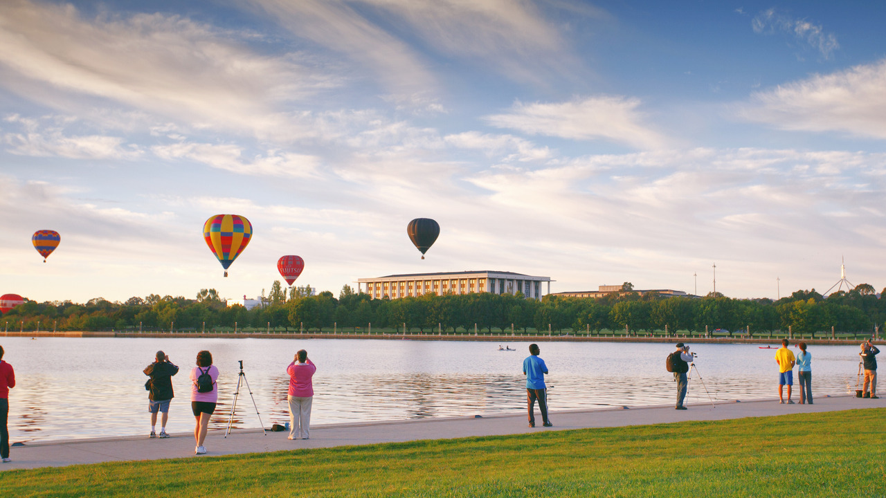 People watch hot air balloons float over a lake. 