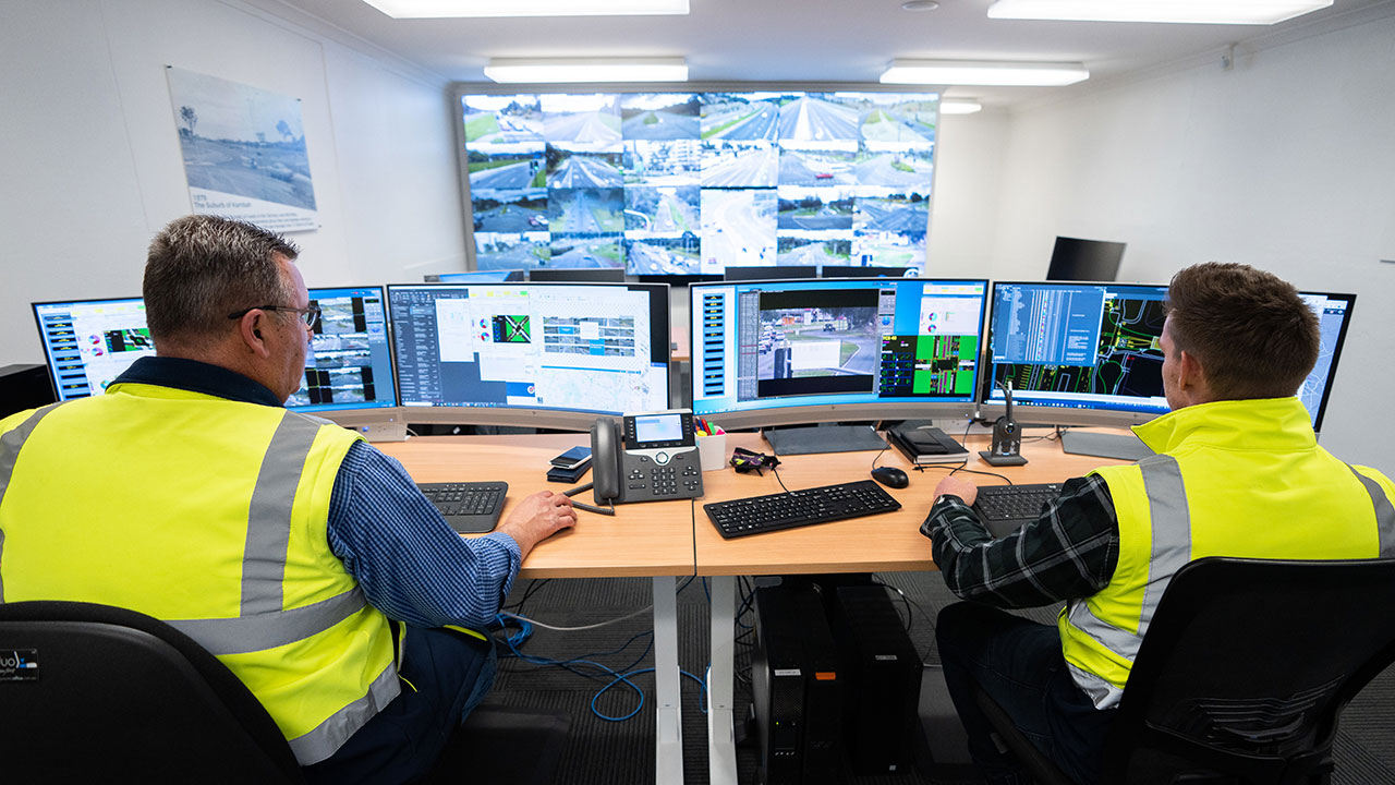 Two men in high-vis vests photographed from behind. They watch traffic on a lot of computers and screens. 