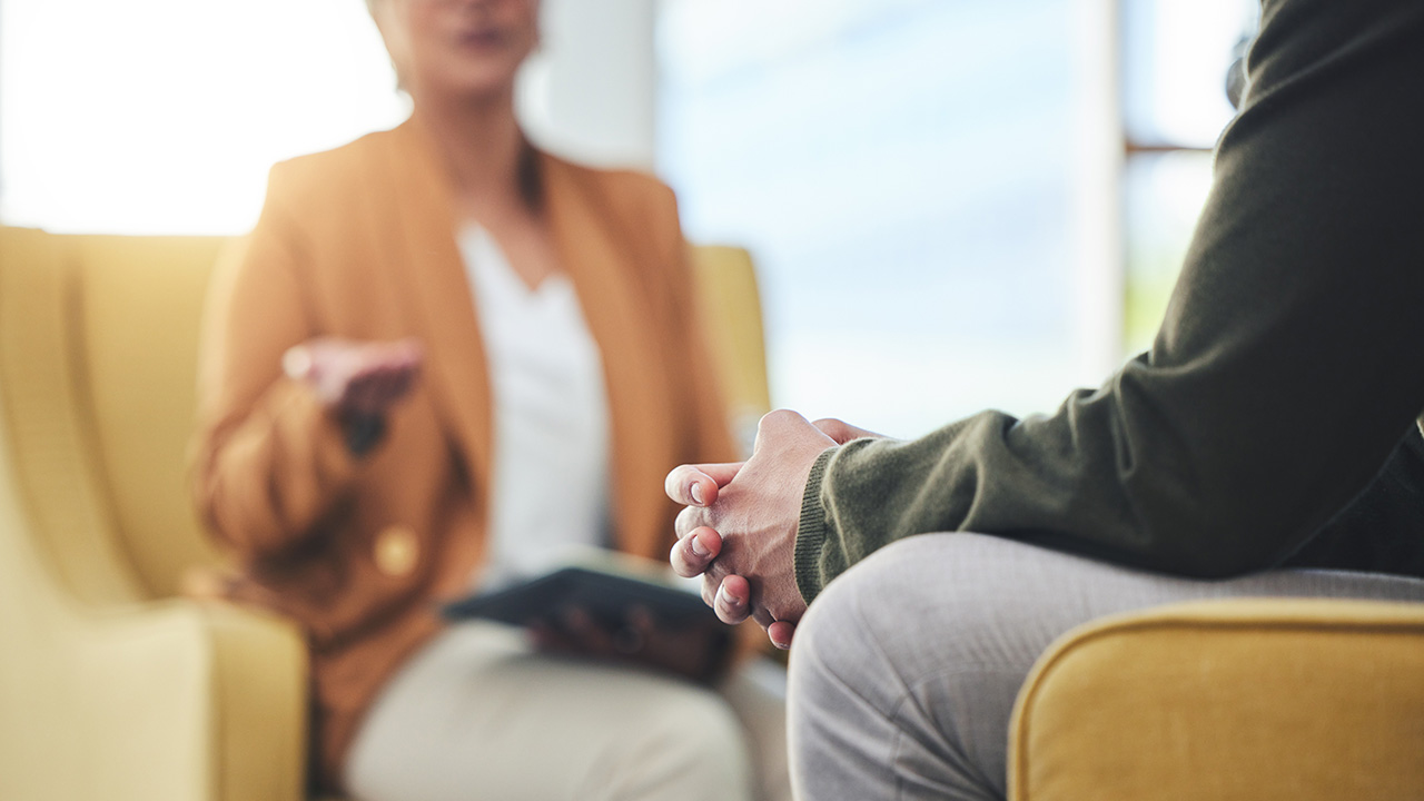 Two people sit in armchairs. One person clasps their hands.