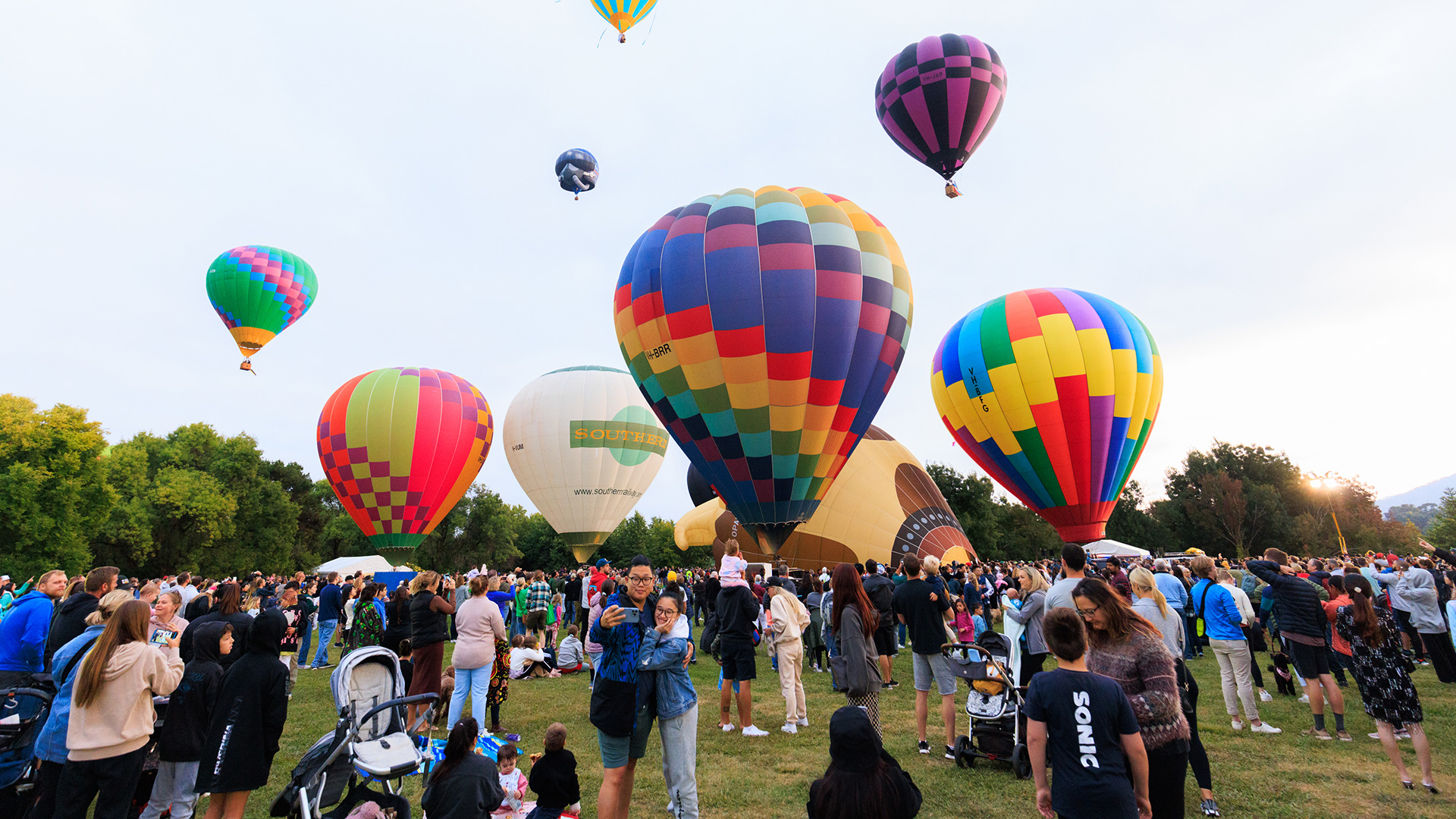 A large crowd stands around looking at a range of colourful hot air balloons.
