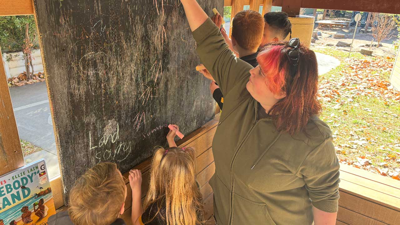 A woman with pink hair reaches up to a blackboard while students stand around her.