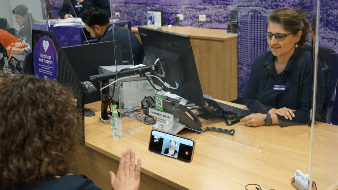 A person being served by an Access Canberra team member. A phone on the table has someone signing Auslan.