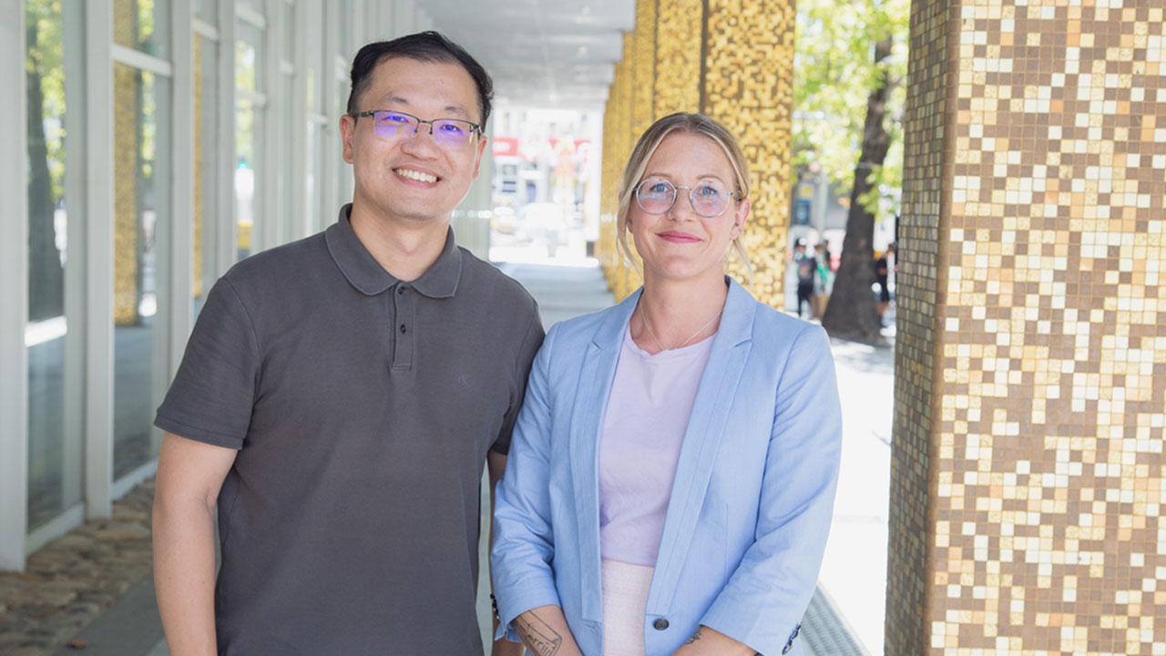 A man and a woman stand outside, smiling. They both wear glasses.