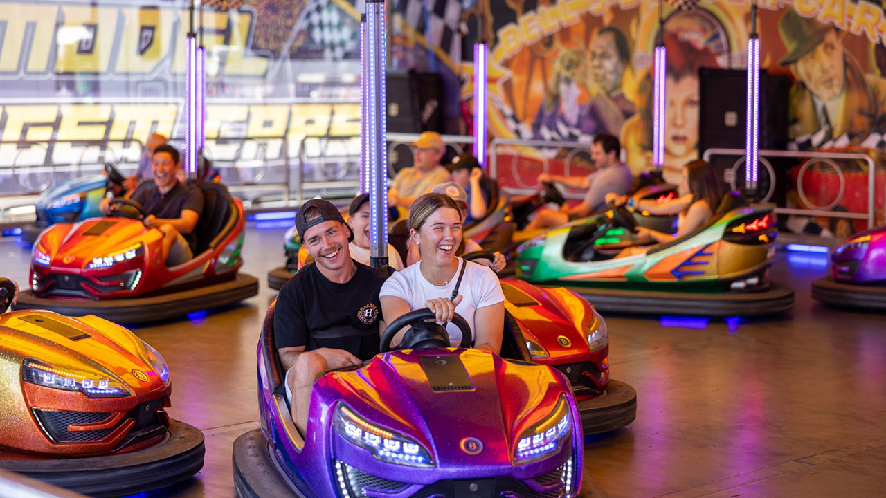 Two people ride in a dodgem car at a carnival, surrounded by others. They are smiling.