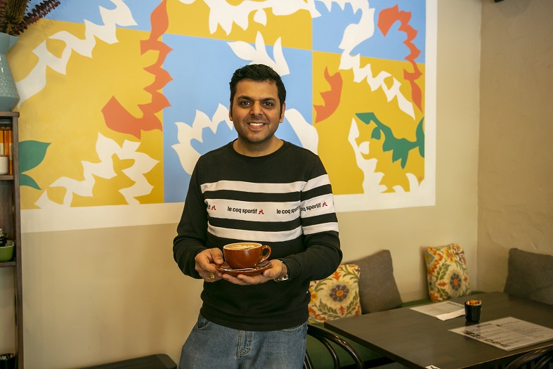 A man, Joy Thakkar, sits at a table holding a coffee in a cup and saucer. There is a bright painting behind him.